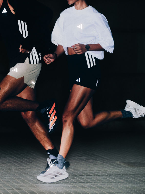 The image shows two people wearing Adidas sportswear, with one person's legs and feet visible in the foreground against a dark background.