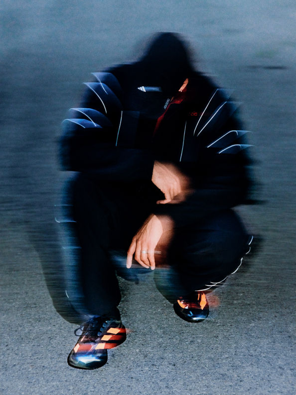 A person in a dark, hooded jacket crouches on a dimly lit road, their face obscured by the shadows.