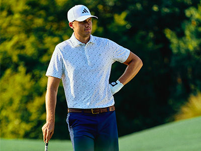 A man wearing a white golf shirt and cap stands on a lush, green golf course surrounded by trees.