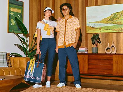 Two individuals, a woman wearing a white shirt and a man wearing a patterned shirt, stand in a room with wooden walls and furniture, including a painting on the wall.