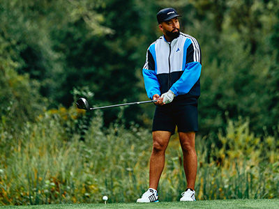 A man with a beard wearing a blue and white athletic jacket is standing on a grassy pitch holding a golf club, with a lush green background of trees and vegetation.