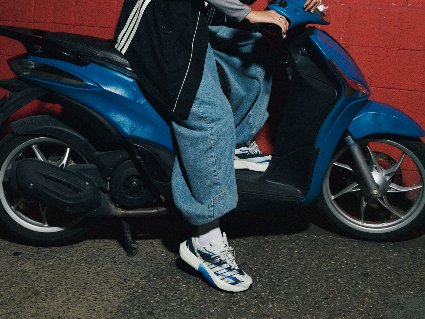 A blue scooter parked on a red-walled background, with a person's legs and feet visible, wearing blue jeans and white sneakers.