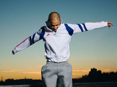 A person wearing a white and blue athletic jacket stands with arms outstretched against a vibrant sunset sky, with a cityscape visible in the background.