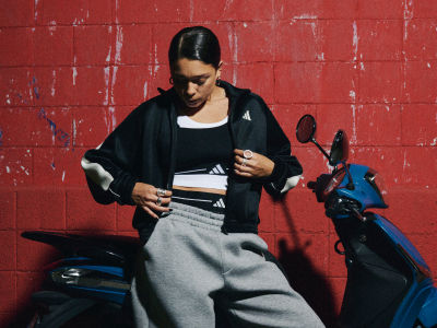 A young woman in a black jacket stands in front of a vibrant red brick wall, with a motorcycle visible in the background.