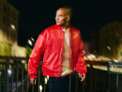 A person wearing a bright red jacket stands in a dimly lit outdoor setting, with blurred structures visible in the background.