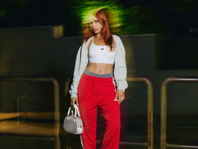 A young woman with long, reddish-brown hair stands in the foreground, wearing a white crop top and bright red pants, holding a small white handbag. The background appears to be a dimly lit outdoor setting, with blurred greenery and structures visible.