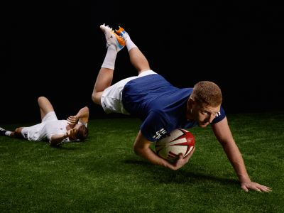 A rugby player in a blue jersey is diving to the ground with the ball, while another player in white is attempting to tackle them on a grassy field.
