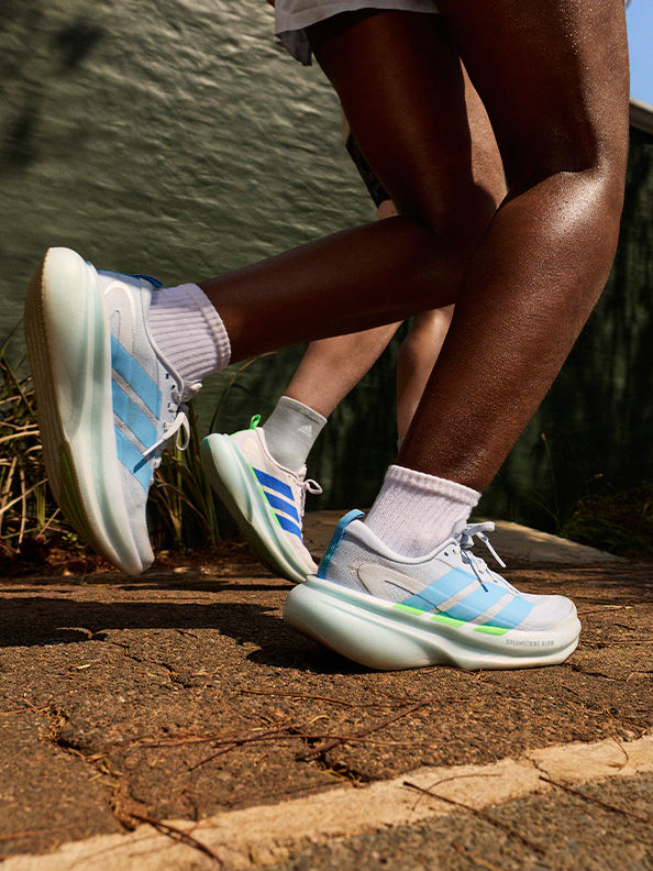 The image shows a person's lower body wearing a pair of colourful, futuristic-looking trainers standing on a wooden surface against a blurred natural background.