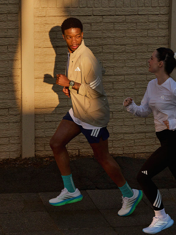 Das Bild zeigt einen jungen Mann, der eine beige Jacke, Shorts und farbenfrohe Sneaker trägt, vor einer Ziegelwand steht und sich einer körperlichen Aktivität oder Bewegung hingibt.