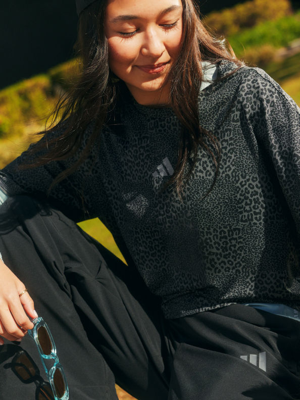 Una mujer joven con pelo largo y oscuro está sentada al aire libre, con una sudadera negra y gris estampada y sonriendo contenta.