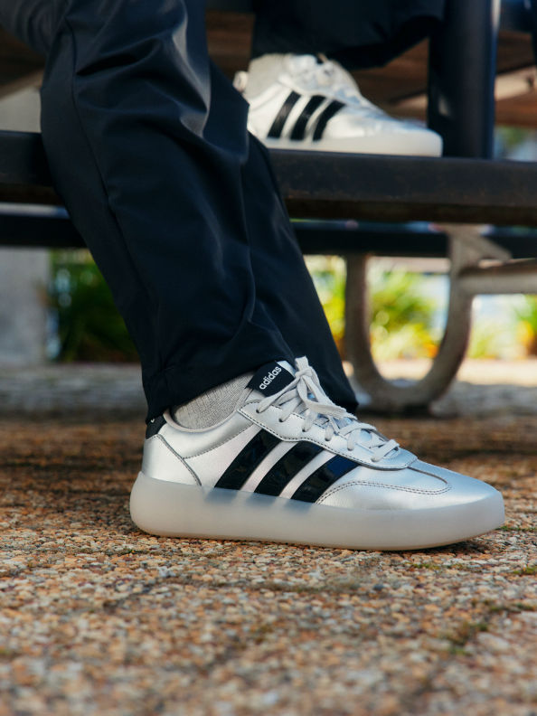 Unas zapatillas adidas blancas y negras sobre los pies de una persona, con un banco de madera y vegetación visible en el fondo.