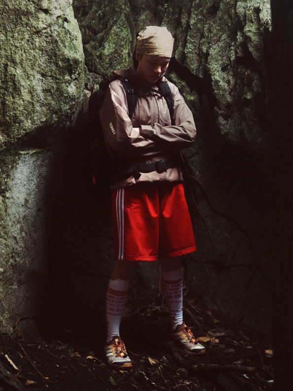 A person wearing a hooded jacket and red shorts stands in a dark, rocky environment surrounded by lush vegetation.