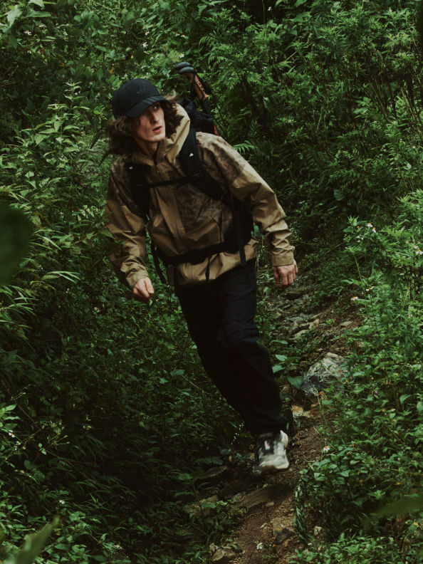A person wearing a camouflage jacket and hat is walking through a dense, lush forest.