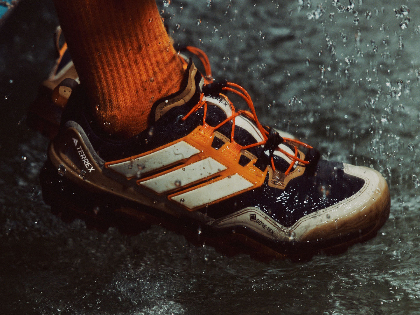 Primo piano del piede di una persona che indossa un paio di scarponi da hiking arancioni e bianchi, con goccioline d'acqua visibili in superficie, posizionate su uno sfondo sfocato e scuro.