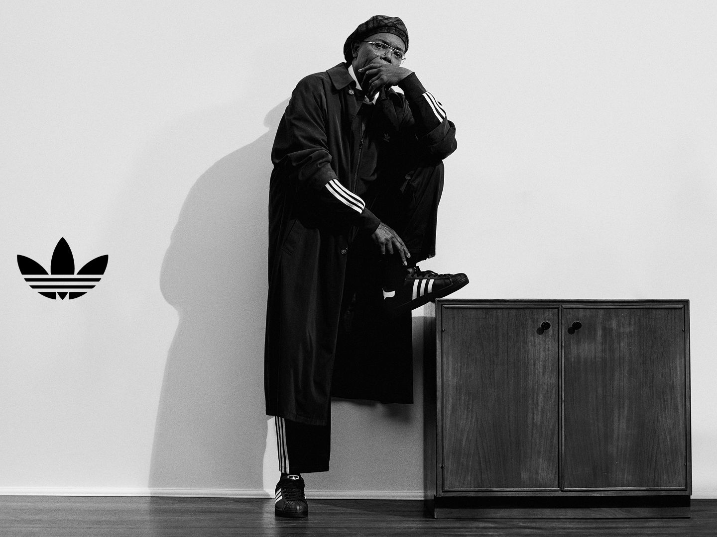Samuel L Jackson con un abrigo largo adidas con mangas de las tres bandas y zapatillas adidas Superstar negras posa con un pie sobre una cajonera de madera, con la mano apoyada en la cara.