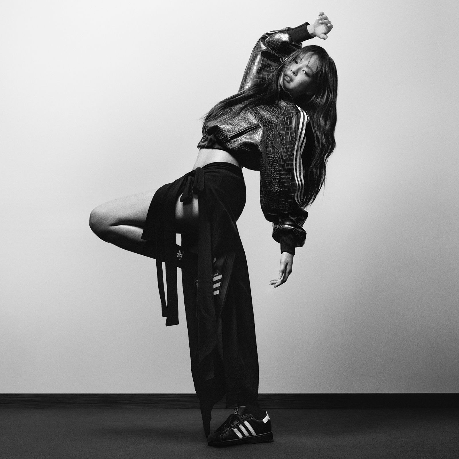JENNIE med teksturert adidas-jakke og svarte adidas Superstar-sko poserer på det ene benet med en hevet arm.