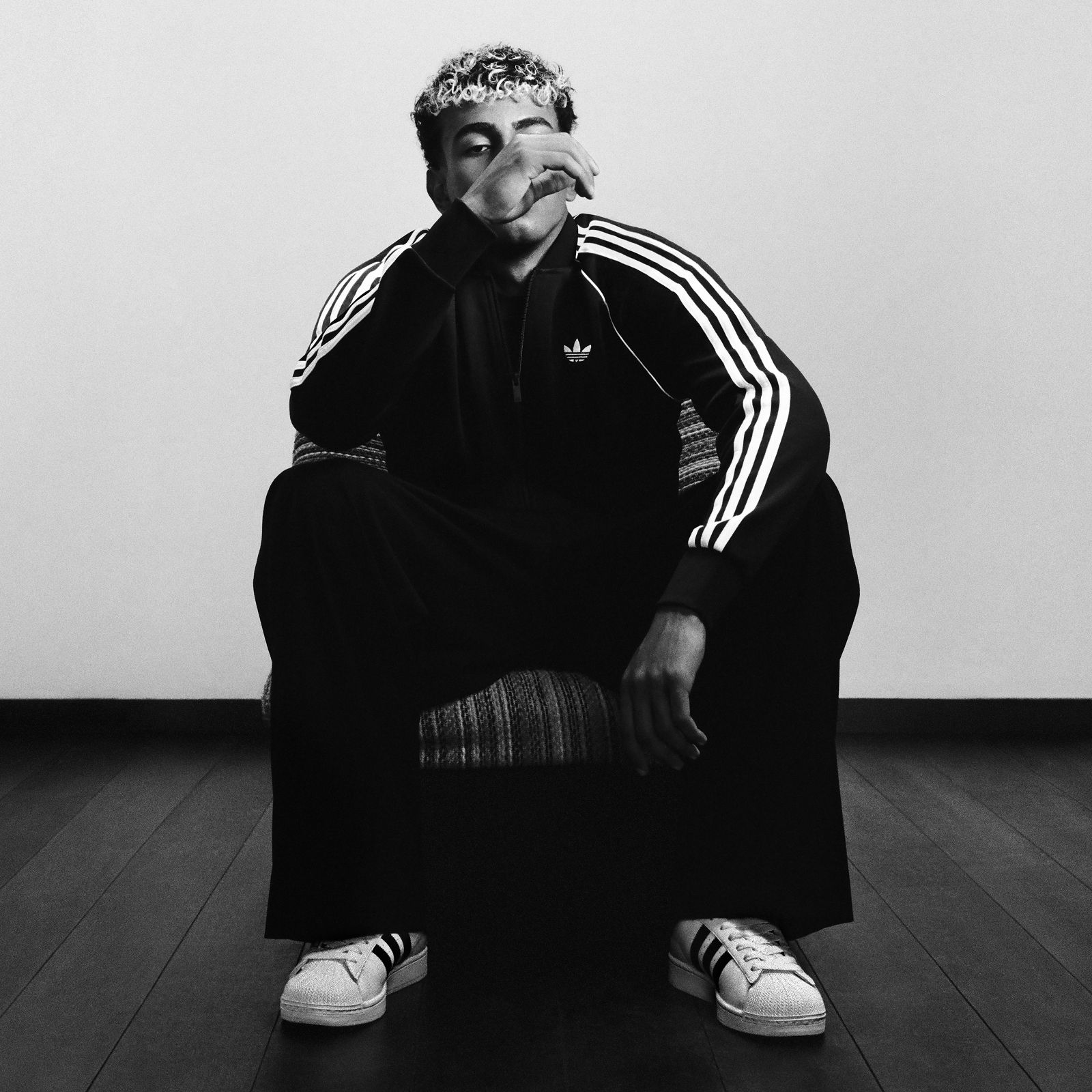 Lamine Yamal luce una sudadera negra de adidas con el logo del Trifolio y zapatillas adidas Superstar blancas y está sentado en un sillón con una mano levantada cerca de su cara.