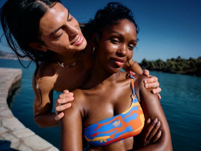 Dos mujeres con trajes de baño se dan un abrazo en una playa con un paisaje de fondo de agua y montañas.