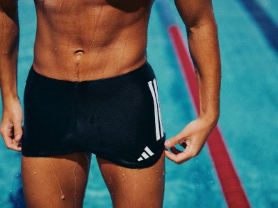 A muscular, shirtless person wearing black athletic shorts stands in front of a vibrant blue background, holding a red exercise band.