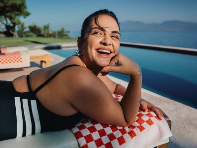 Una mujer sonriente con traje de baño sentada junto a una piscina con vistas panorámicas al océano de fondo.