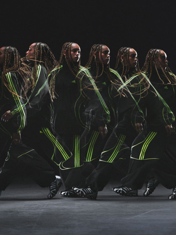 Un grupo de bailarines con disfraces negros actúa en un escenario oscuro, sus movimientos crean senderos dinámicos en luz verde que acentúan su coreografía.