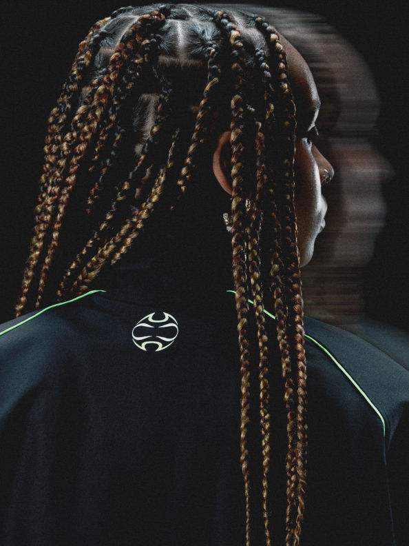 La imagen muestra a una persona con un intrincado peinado trenzado sobre un fondo oscuro, con un logo visible en su ropa.