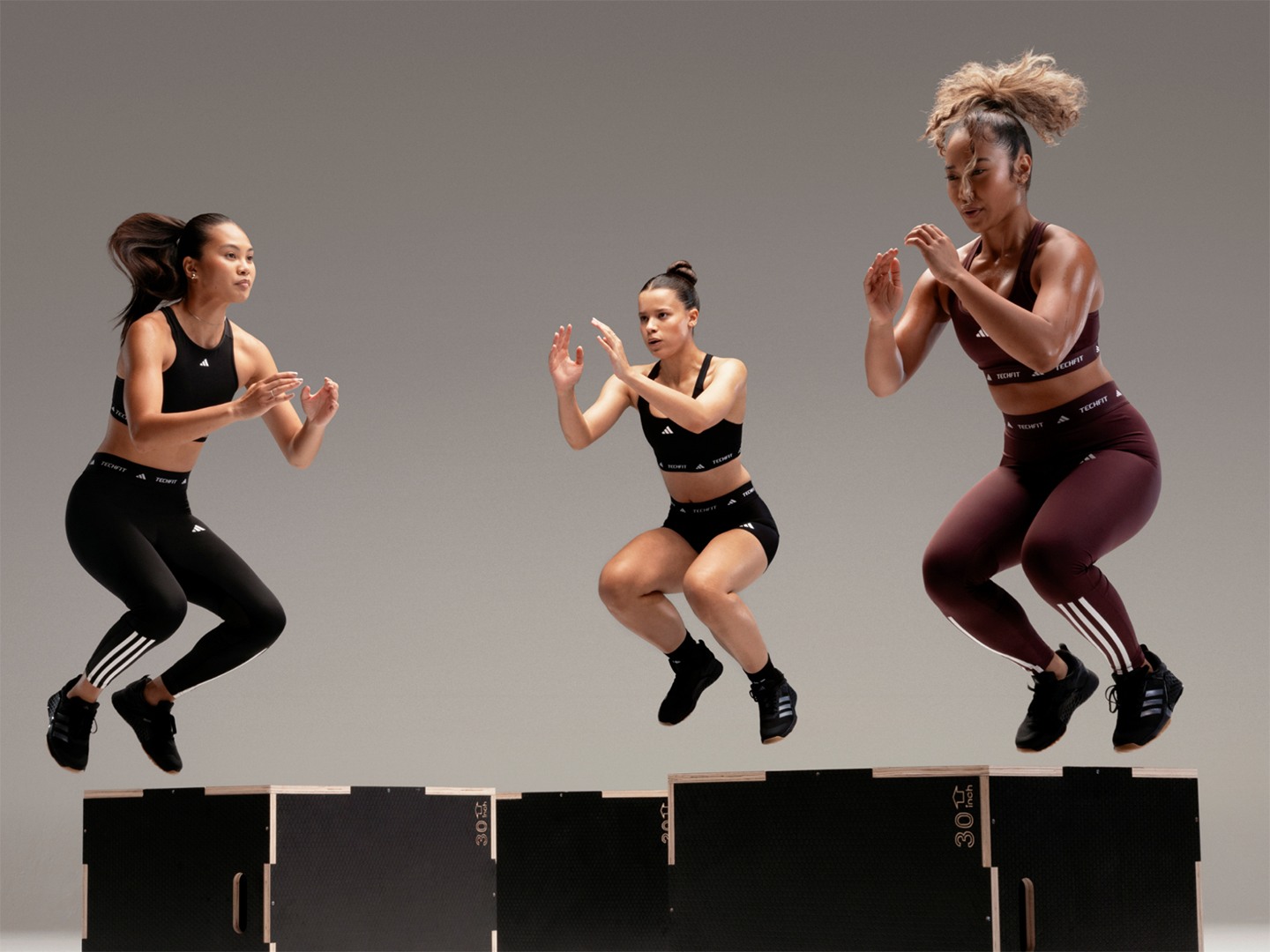Three women in athletic wear are performing synchronized dance moves on a set of black platforms against a plain background.