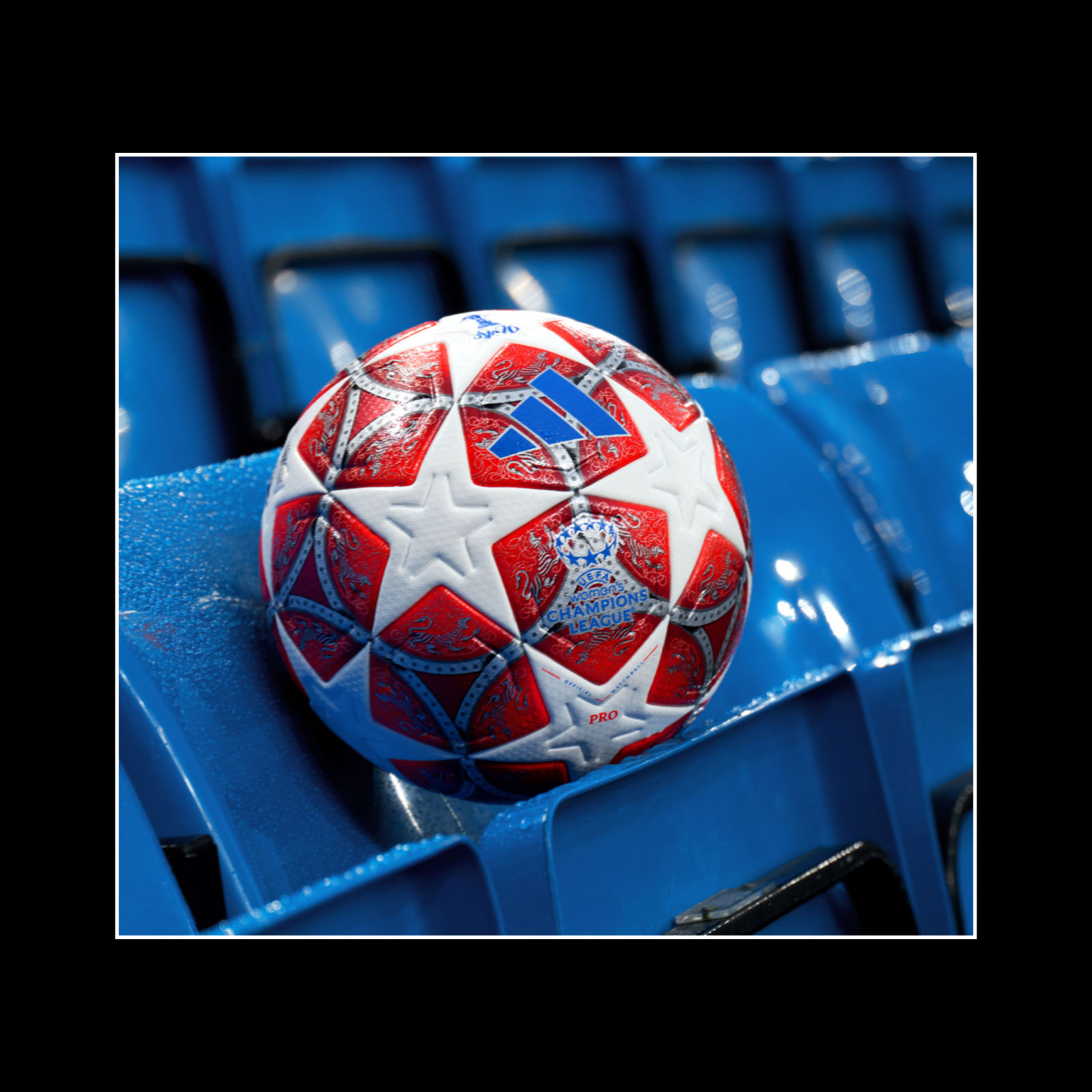 A vibrant, star-patterned soccer ball rests on a blue stadium seat, with the blurred background suggesting a sports arena or stadium setting.