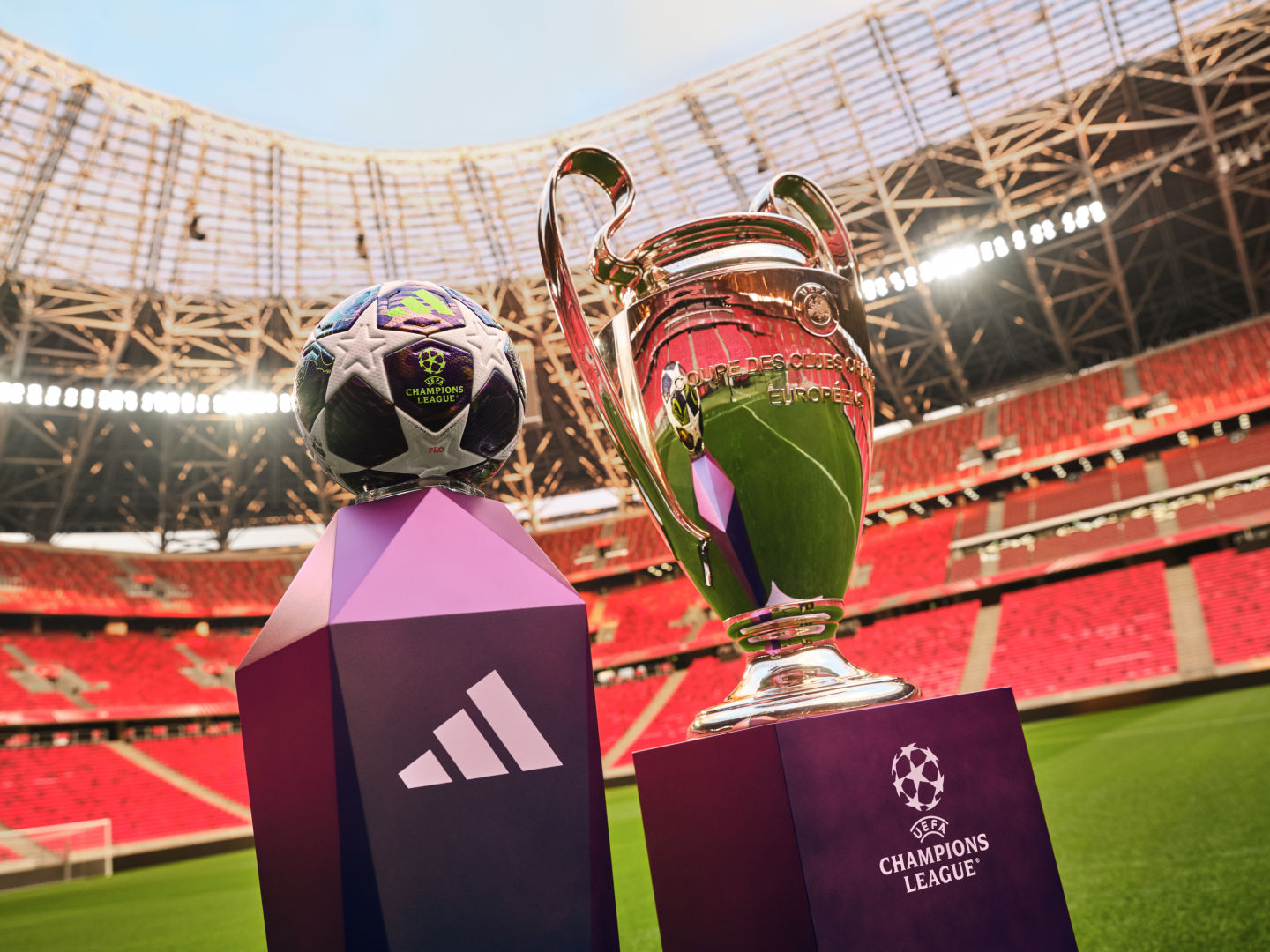 The image shows the UEFA Champions League trophy and a soccer ball on a field in a large stadium.