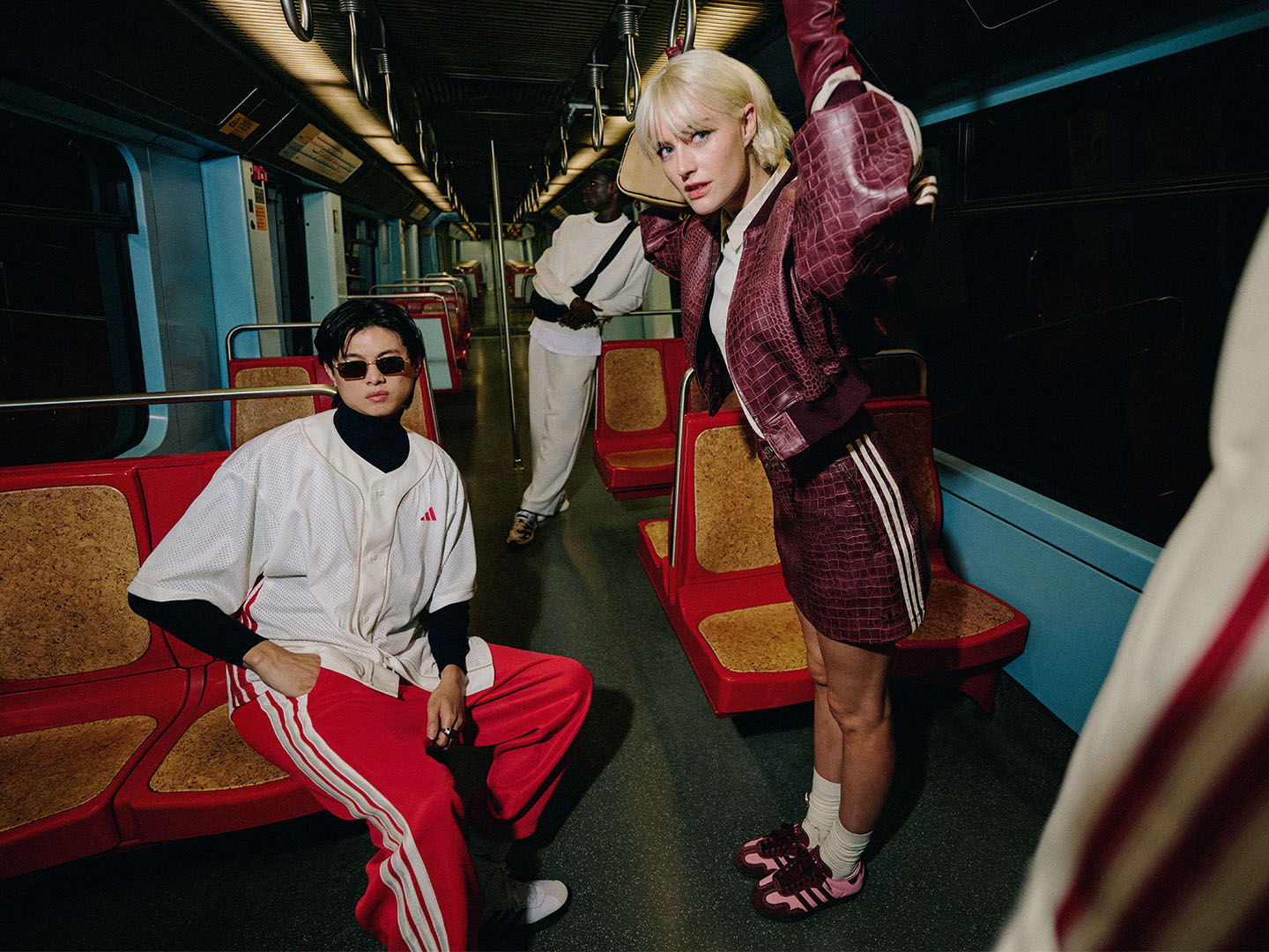 Two individuals, a woman with blonde hair and a woman with dark hair, are standing in a colourful, retro-style train car. The background features vibrant red and yellow seats, adding to the visually striking atmosphere.