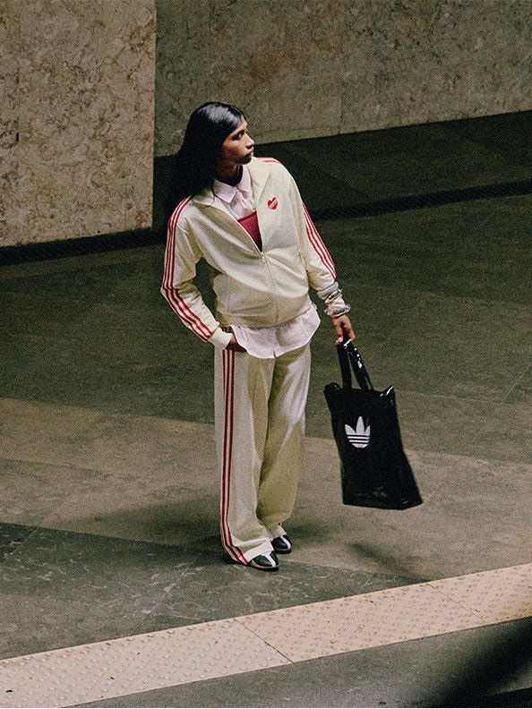 白とピンクのトラックスーツを着て、薄暗い屋内で、adidasのロゴが入った黒いバッグを持っている人物の姿がみえる。