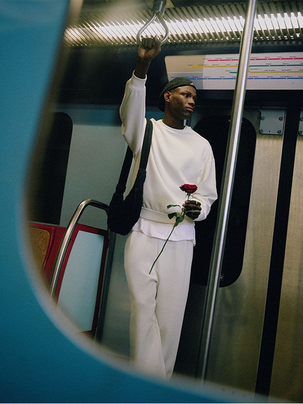 赤いバラを着た白い服を着た人物が、地下鉄の車内に立っている。背景には列車の車内が映っている。