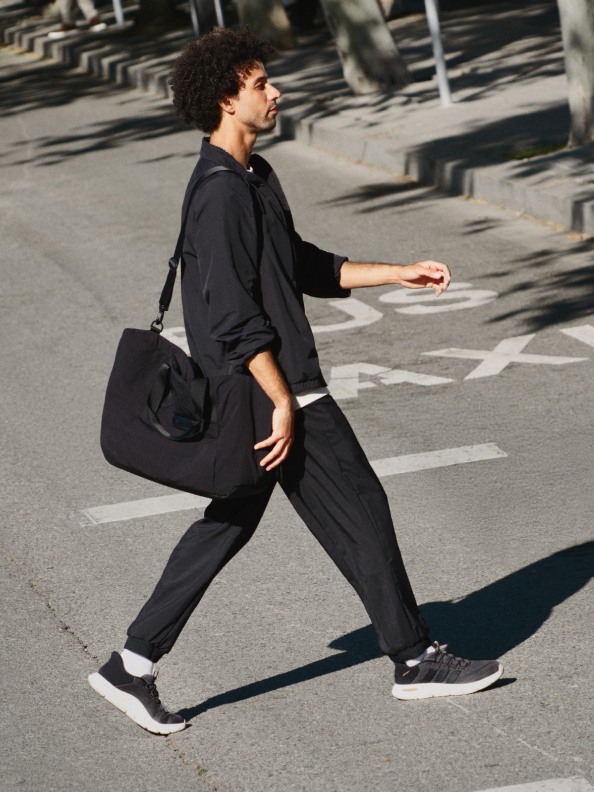 A person in a black outfit carrying a bag is walking on a city street with rows of parked cars in the background.
