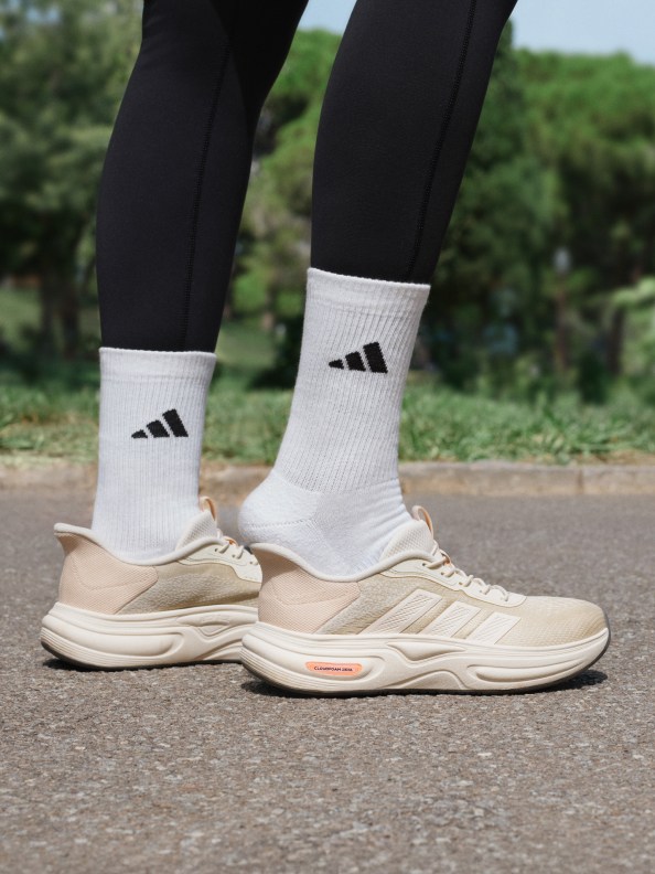 The image shows a pair of white Adidas socks and beige athletic shoes on a person's feet, standing on a paved road surrounded by lush greenery in the background.