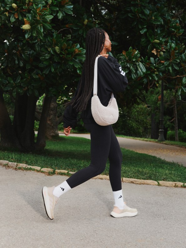 A person wearing black clothing and white sneakers is walking on a paved path surrounded by lush greenery and trees.