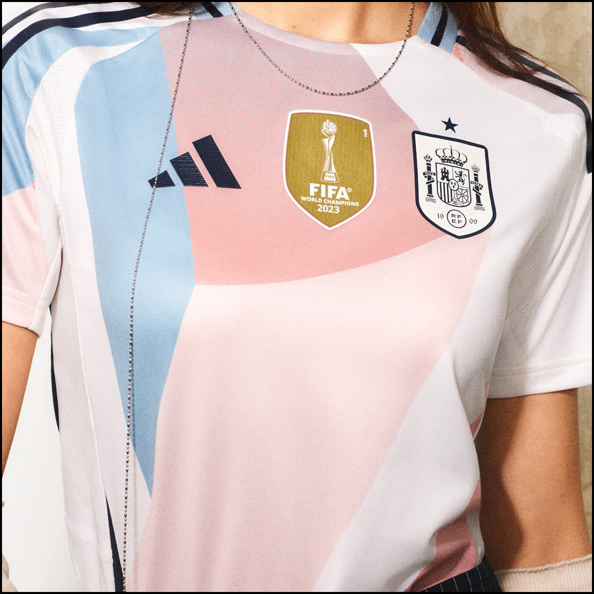 Ein weiß-rosa Trikot mit dem FIFA 2022 Logo und dem Logo der spanischen Nationalelf, das von einer Person im Vordergrund getragen wird.