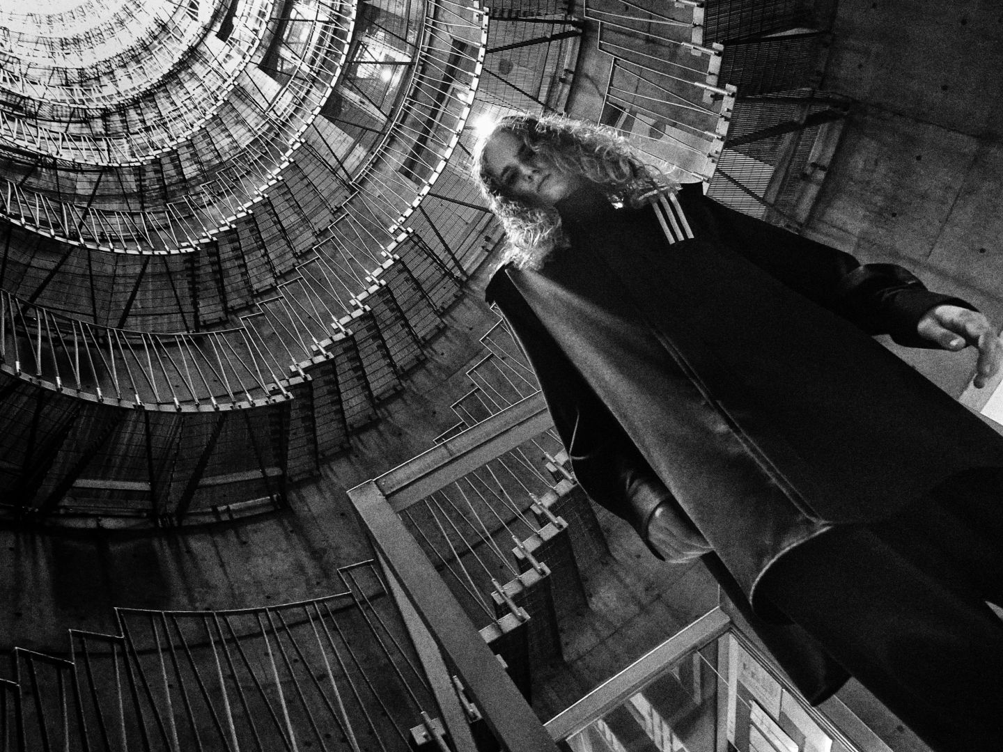 A lone figure in a dark coat stands amidst a towering, intricate structure of metal and glass, creating a striking contrast between the human presence and the architectural grandeur surrounding it.