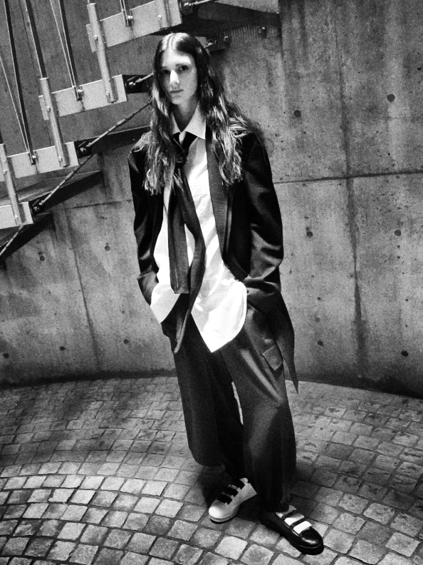 A person wearing a long black coat and holding a white object stands in front of a brick wall, with a tiled floor visible in the foreground.