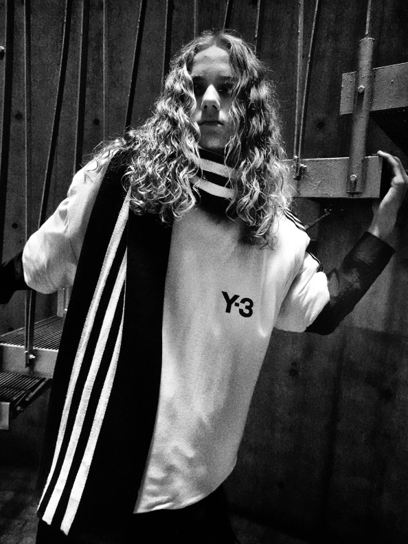 A person with curly hair wearing a white shirt with the Y3 logo stands in front of a dark, wooden background.