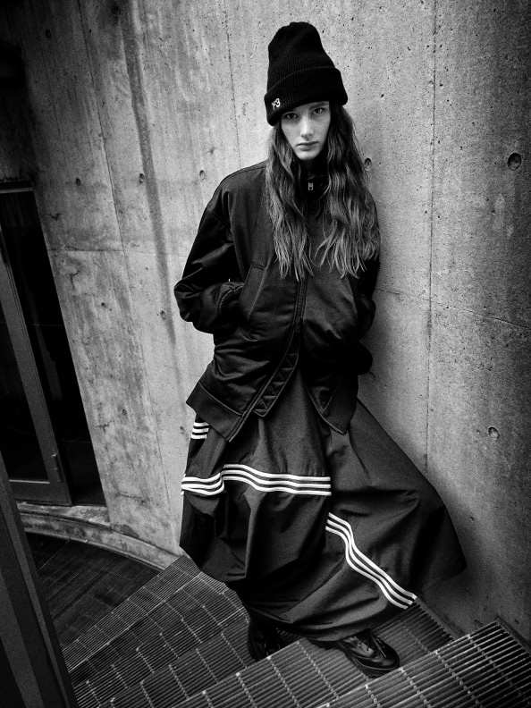 A person wearing a black jacket and hat stands against a concrete wall, their long hair visible as they gaze downward.