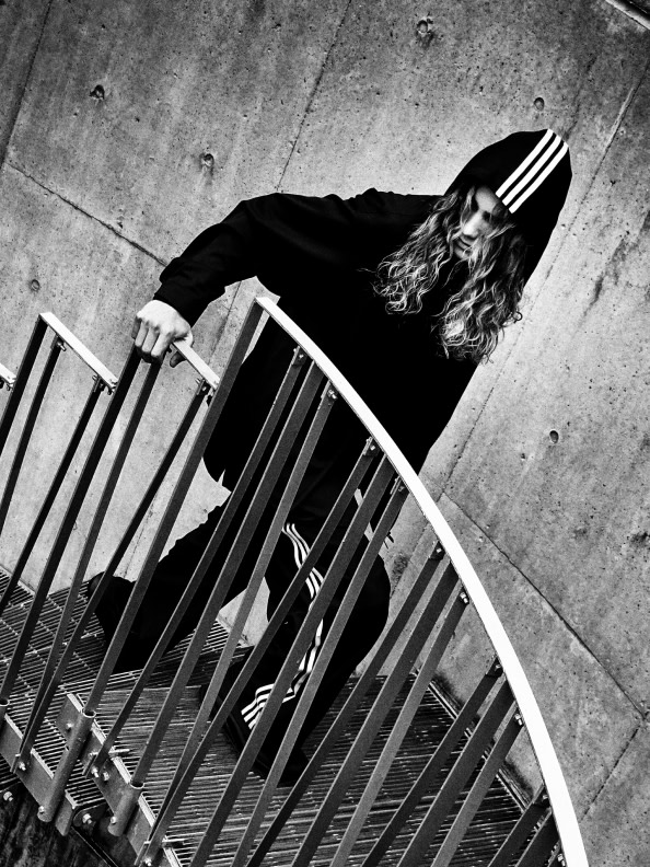 A person with curly hair reclining on a metal chair against a concrete wall.