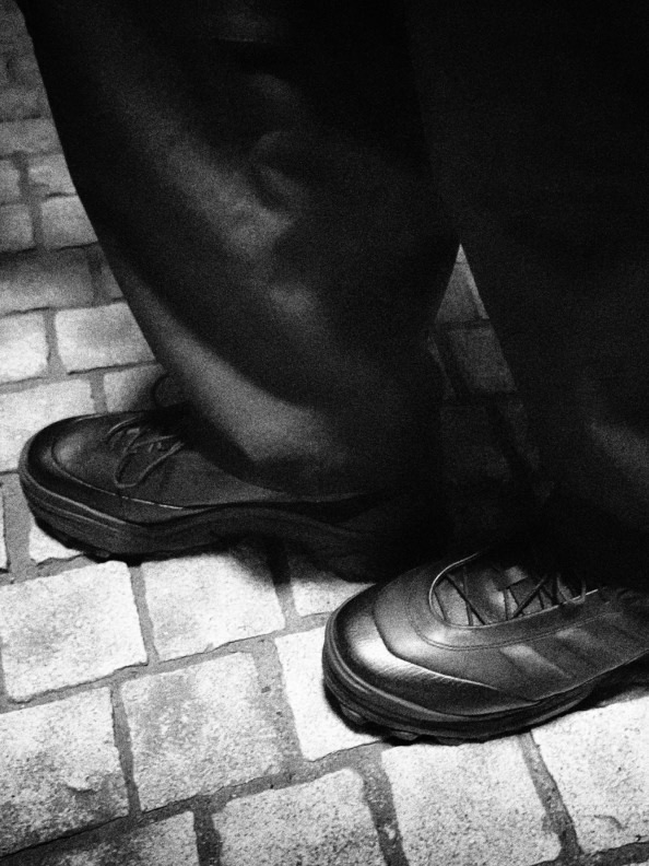 A pair of black leather shoes rests on a tiled floor, with the lower legs and feet of the person wearing them visible in the foreground.