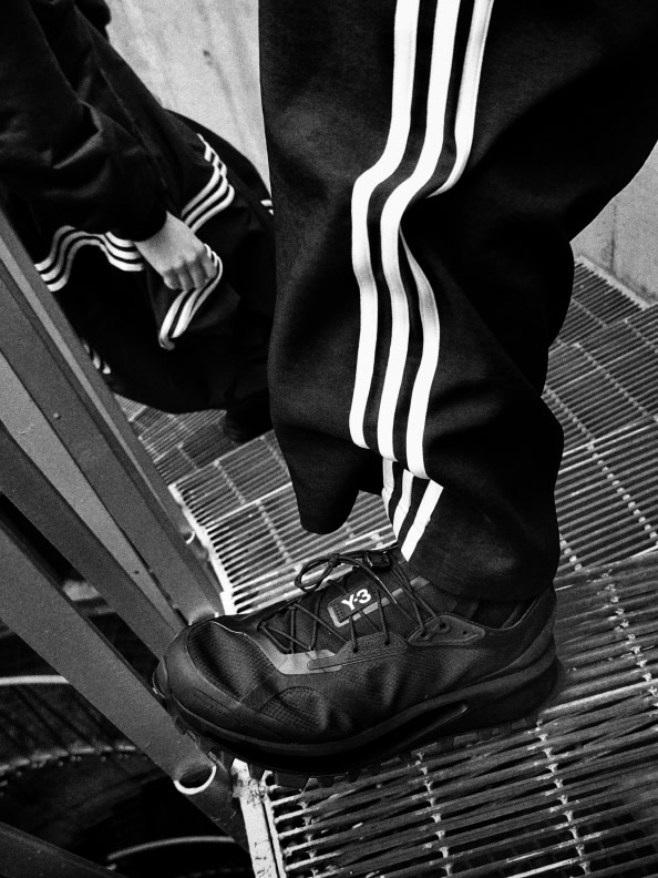 The image shows a close-up view of a person's legs wearing black-and-white striped athletic pants and black sneakers standing on a metal grate.