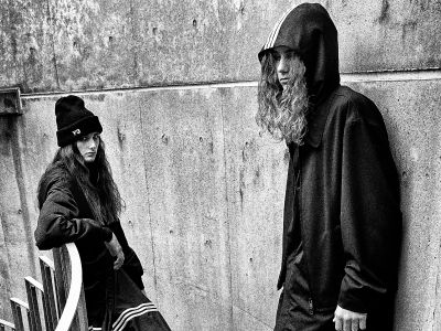 Two individuals wearing dark hooded cloaks stand in front of a concrete wall, creating an ominous and mysterious atmosphere.