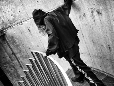 A person in a dark jacket stands in front of a concrete wall, with a graphic design or pattern visible in the background.