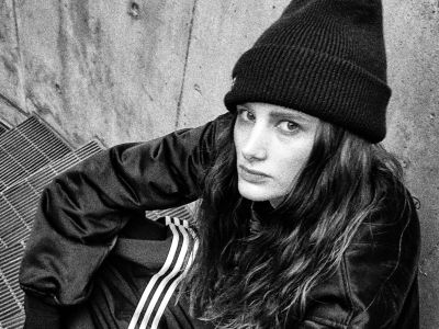 A person wearing a black beanie and jacket stands in front of a weathered, wooden wall.