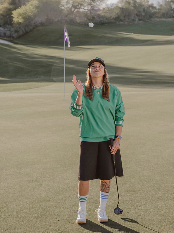 A young woman in a green sweatshirt and black shorts stands on a golf course, with a golf club in her hand and a flag in the background.