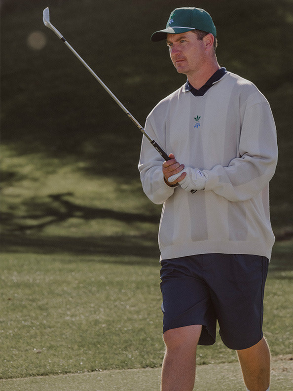 A man in a baseball cap and casual clothing stands on a grassy golf course, holding a golf club and preparing to take a swing.