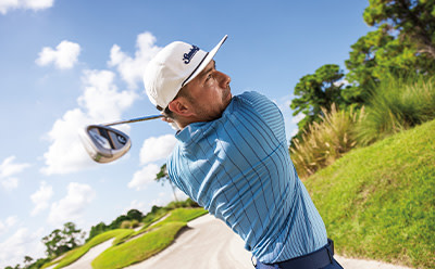 A golfer in a blue striped shirt and white hat is swinging a golf club on a lush green golf course with trees and clouds in the background.