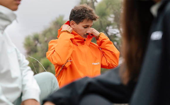 A person wearing a bright orange jacket stands in the foreground, while a group of people can be seen in the background.